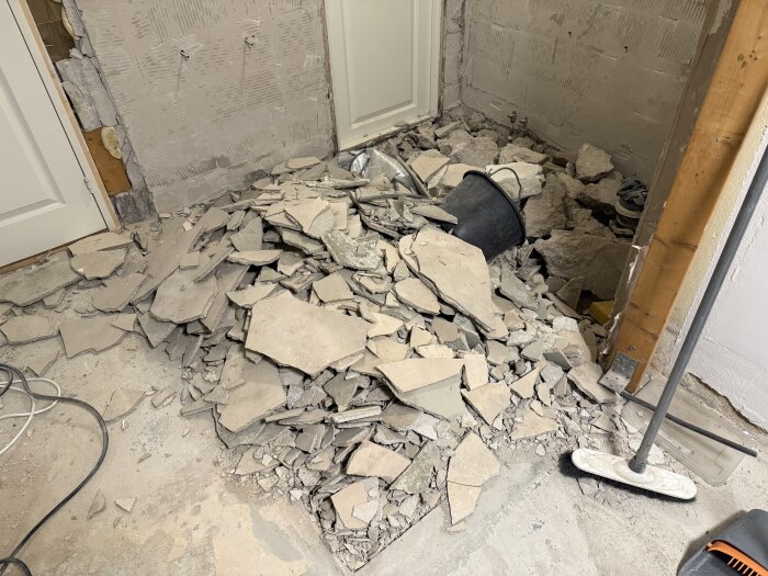 Bitar av sprucket flytspackel och betong på ett golv, en svart hink syns bland rasmassorna, samt en borste lutad mot en vägg.