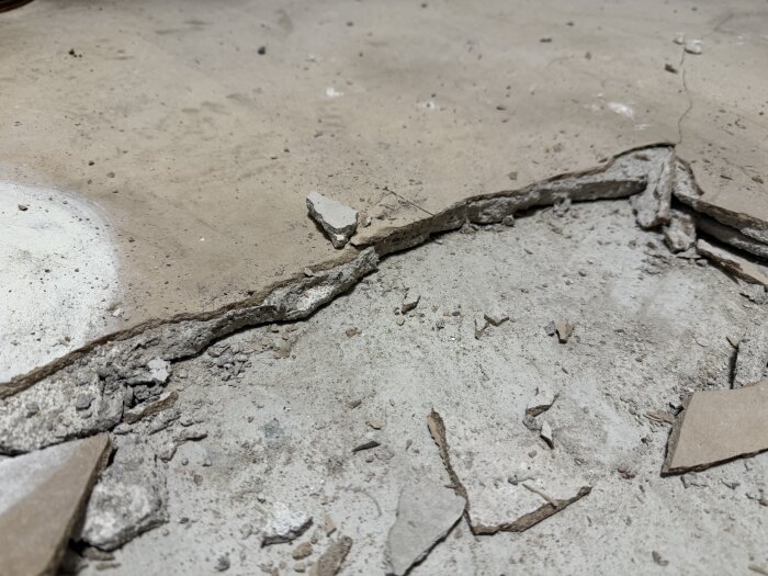 Sprucket flytspackel på ett betonggolv. Spacklet har lossnat i bitar och ligger omkring öppna sprickor i underlaget som visar den ursprungliga betongen.
