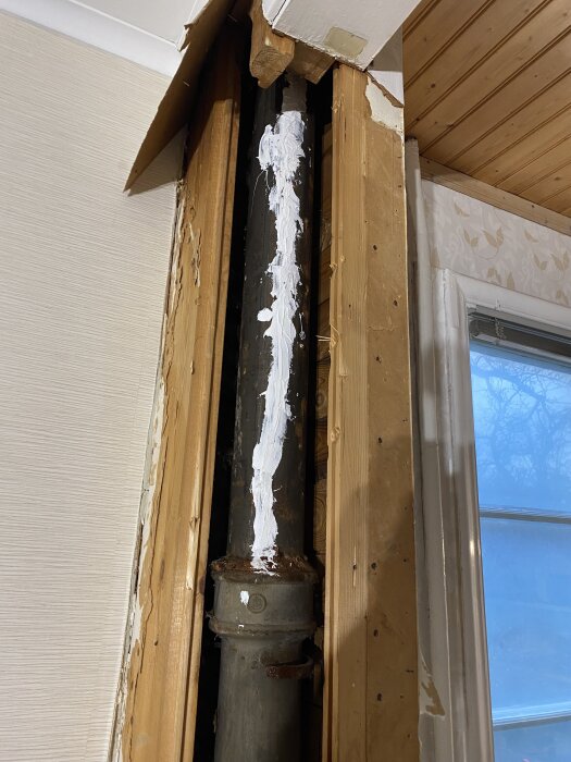 Gammalt avloppsrör med sprickor och vit tätning, omgivet av avskalade trä- och gipsskivor. Renovationsarbete i husvägg.