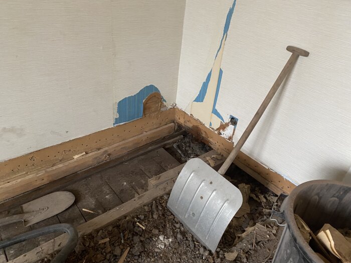 Rivningsprojekt med trasigt avlopp; golv och vägg delvis öppnade. Spade och skovel ligger på golvet, skräp och isolering i en hink bredvid.