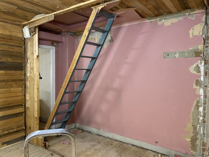 En rosa vägg med delvis borttagen tapet, en blå stege och blottat trä i ett rum under renovering.