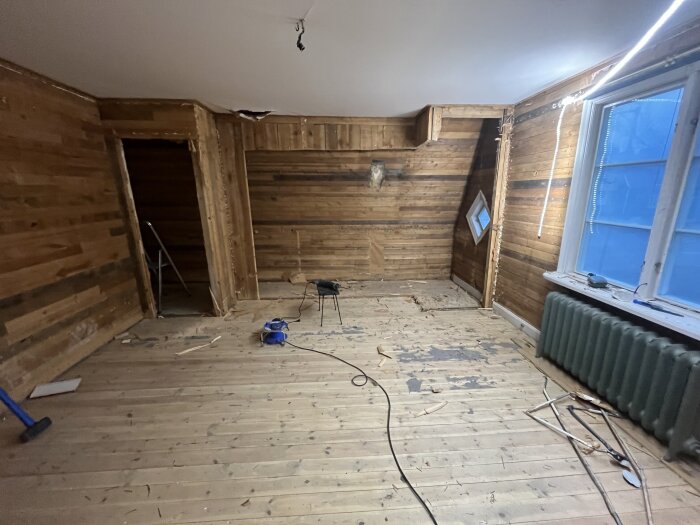 Övervåning under renovering med avklädda träväggar och golv, ett löst fönster, ett element under fönstret och verktyg på golvet.