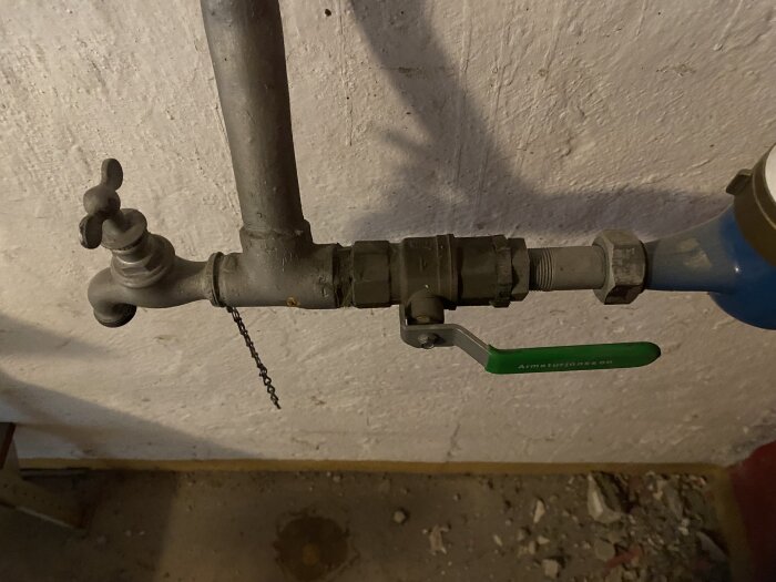 Gammal vattenventil av metall med grön handtag på ett rör, bakgrund av vit vägg.