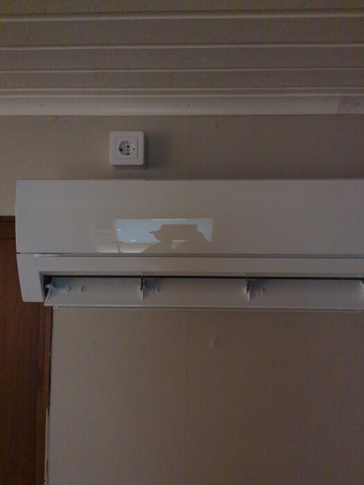 Vit luftkonditioneringsenhet monterad på vägg under ett eluttag i ett rum med vitmålad träpanel i taket.