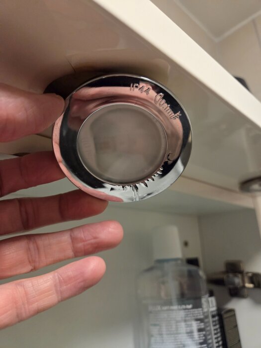 Rund badrumslampa under skåp med texten "IP44", en hand visar lampan närbild.