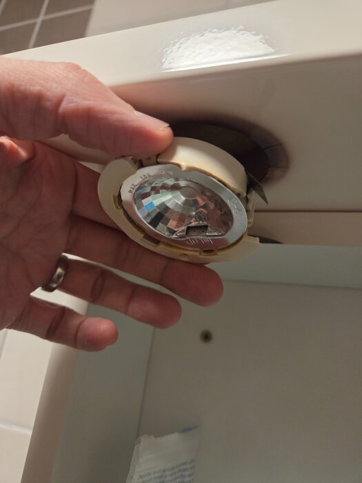 En hand håller en trasig och isärplockad infälld lampa under ett skåp; spår av sprickor ses runt öppningen.