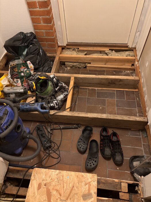 Under golvrenovering med blottade reglar, skor, verktyg och skyddsutrustning på brun kakelgolv, samt en dammsugare och strömkablar.