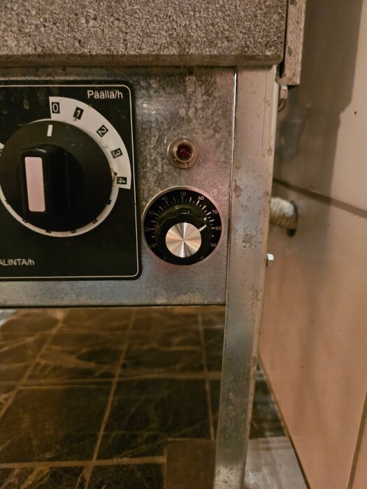 Metallpanel med justerbara vred och en röd LED-lampa. Reglering och tidsinställning för bastuaggregat visas.