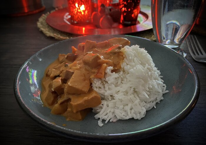 Tallrik med ris och köttgryta i krämig sås, innehållande korv och grönsaker, serverad vid ett bord med tända röda ljus i bakgrunden.