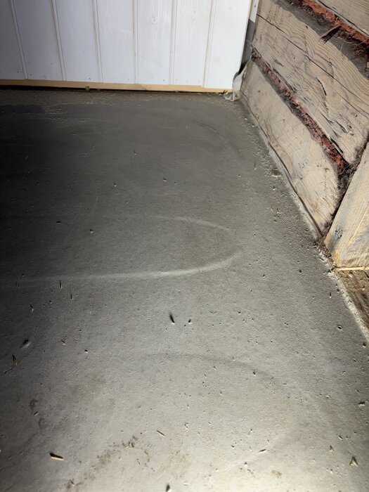 Golv med spackel och synliga sprickor nära vägg; en slang kan anas under ytan. Väggen är delvis av träpanel.