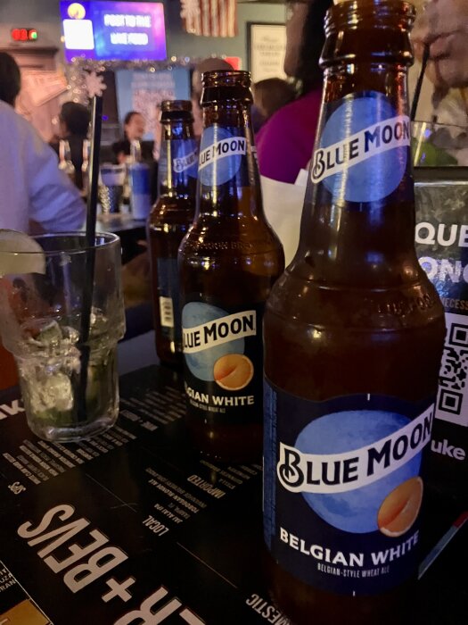 Flaskor av Blue Moon Belgian White öl står på ett bord i en livlig bar med folk och dekorationer i bakgrunden.