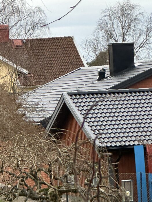 Hus med ny installerat tak, som framstår ojämnt. Byggnaden står i närhet av andra hus, omgiven av träd och buskar.
