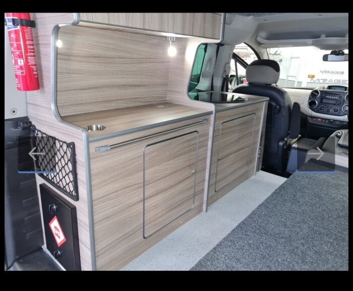 Inredd bil med träpanel, förvaring och belysning inuti skåpbil, med brandsläckare monterad och passagerarstol i bild.