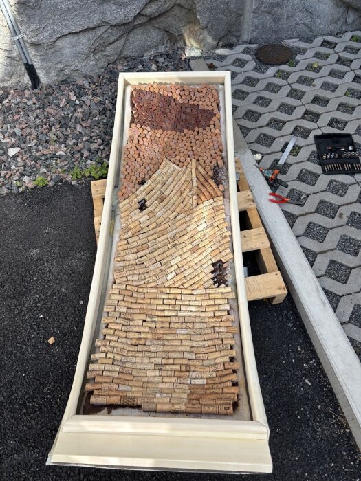 rektangulär dörrkarm fylld med vinkorkar i olika mönster, placerad på pall vid stenkant, verktyg som sågar och borrar syns bredvid