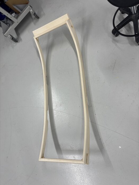 En handgjord träram med böjda kanter ligger på ett golv i en verkstad. Ramen har slipa hörn och är under arbete för ett byggprojekt.