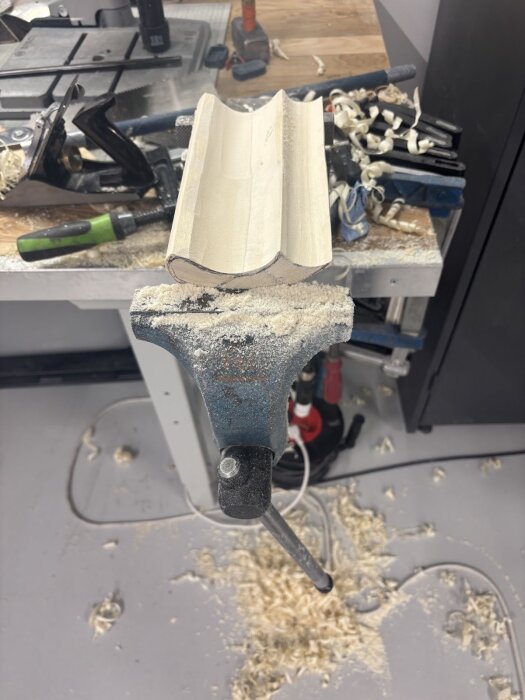En halvfärdig träbit spänd i arbetsbänk omgiven av träspån och verktyg i en verkstad för dörrhandtagsprojekt.