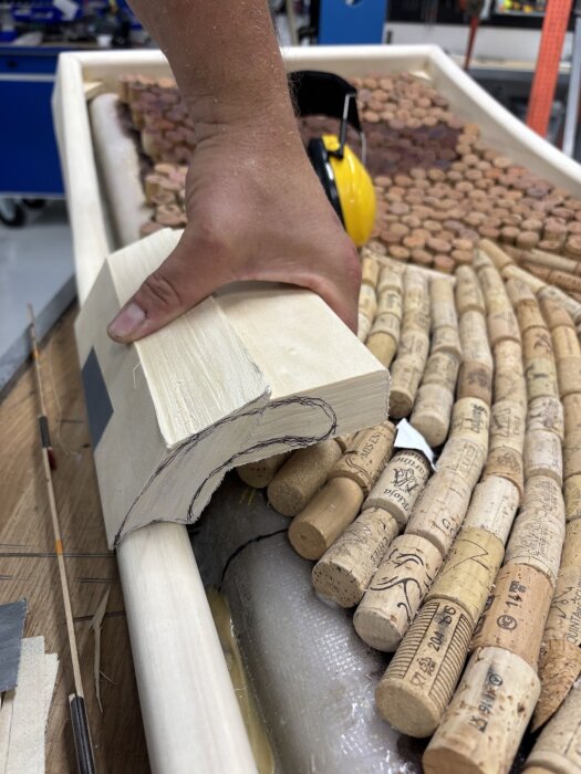 Hand håller träformad mockup mot dörrkarm; korkar och träram i bakgrunden. Arbetsverkstadsmiljö synlig med skyddsutrustning.