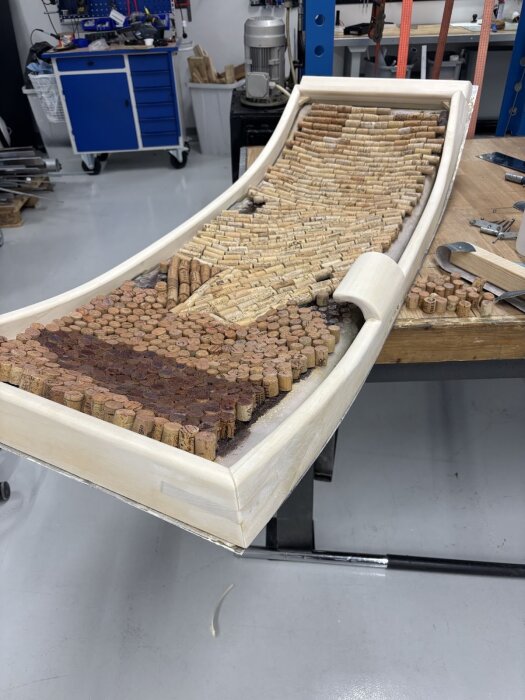 Träram fylld med vinkorkar, placerad på arbetsbänk i verkstad. Ramens kurvor och släta yta syns, samt verktyg och material runt omkring.