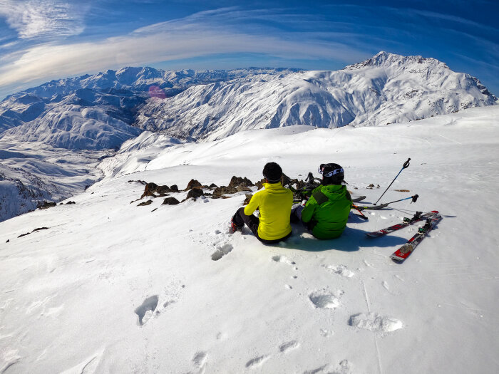 Två personer i skidkläder sitter på en snötäckt bergssluttning med skidor bredvid, och utsikt över snötäckta berg i fjärran.