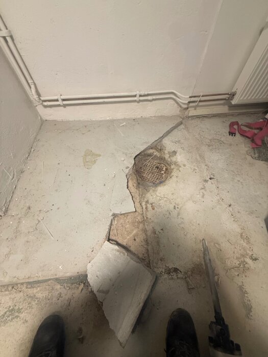 Sprucken betongplatta och ett dräneringshål på golv, med rör längs väggen. Möjligen en tidigare garderobshörna efter borttagen panna.