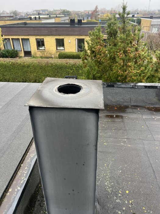 Öppen skorsten utan huv på ett hustak med träd och flera hus synliga i bakgrunden.
