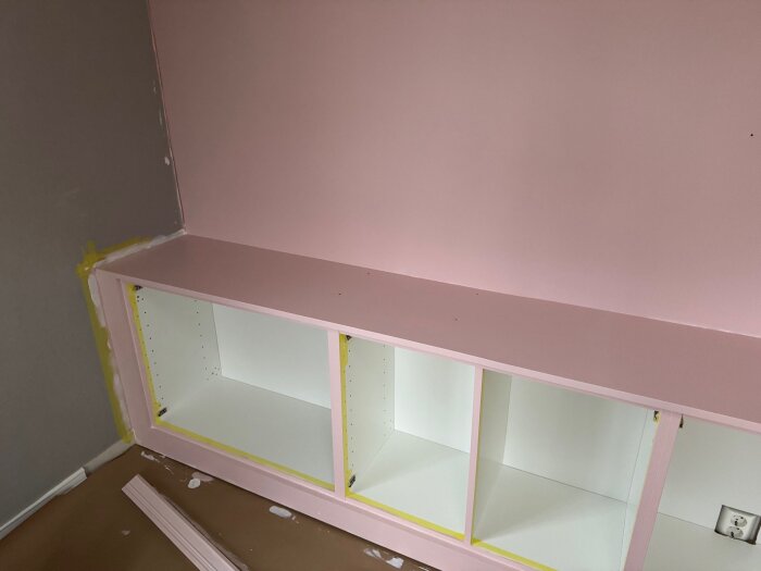 Platsbyggd bokhylla målad i rosa, yta med synliga ojämnheter, tejpade kanter. Utgångspunkten är en fråga om appliceringsteknik och resultat.