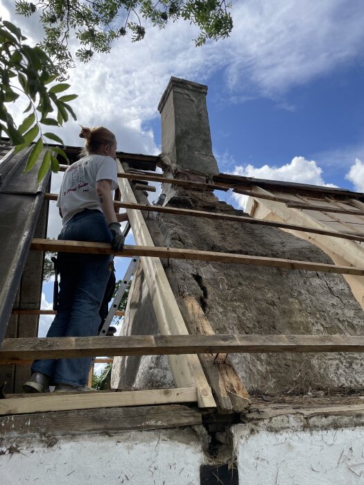 Person inspekterar en gammal skorsten på taket av ett hus under renovering, med trästegar och blå himmel i bakgrunden.