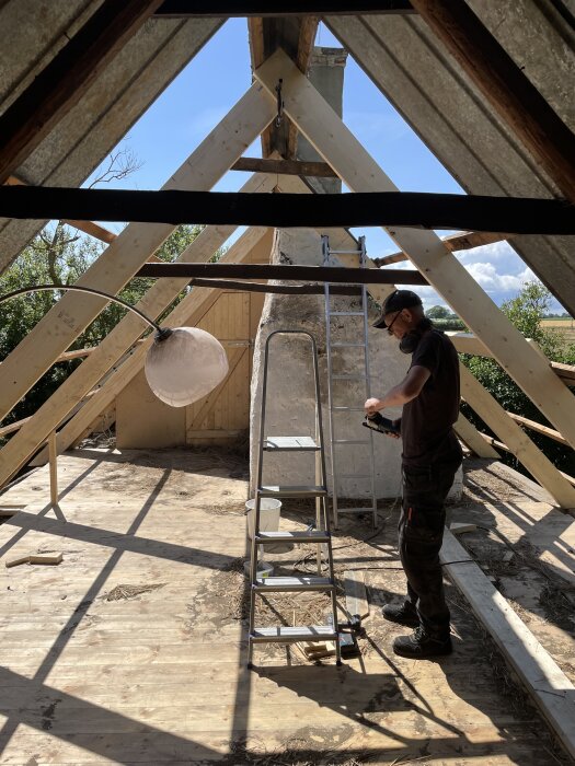 Byggnadsarbetare på vind med gammal skorsten, omgiven av träbjälkar, förbereder renovering; stege och verktyg synliga.