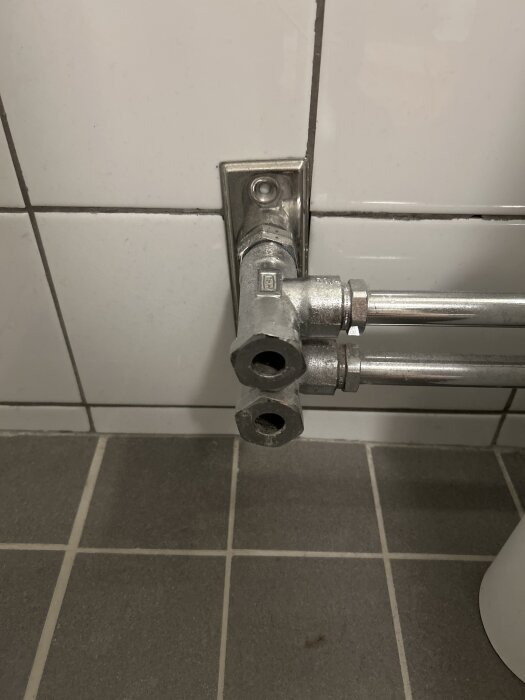 Två metallrör kopplade till en väggfäst anslutning på vit kakelvägg, över grått klinkergolv.