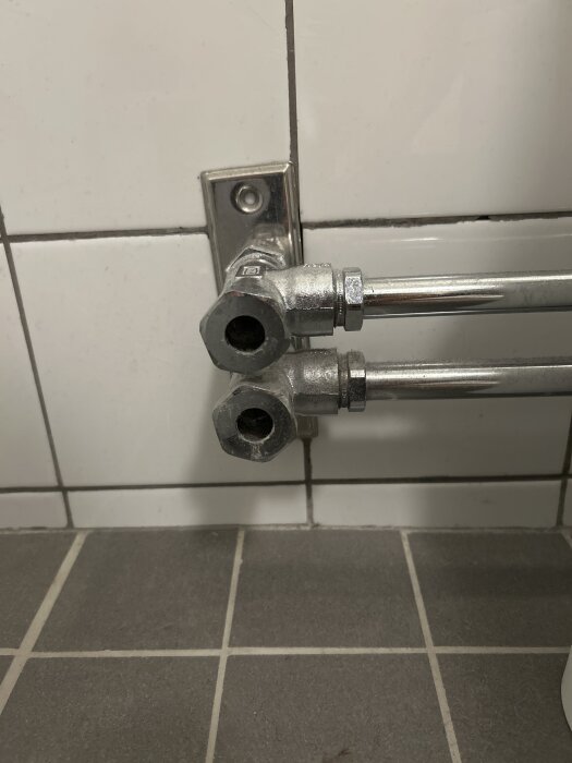Två anslutningsrör av metall monterade på en kaklad vägg i ett badrum, med en grå klinkergolv nedanför.