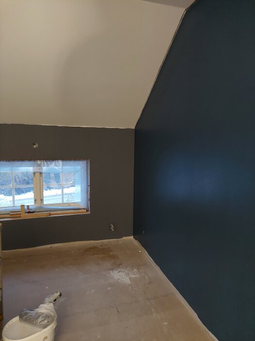 Ett rum under renovering med en målad mörkblå vägg, grå vägg och vitt tak. Ett fönster med vinterlandskap och plasttäckta ytor syns i bakgrunden.