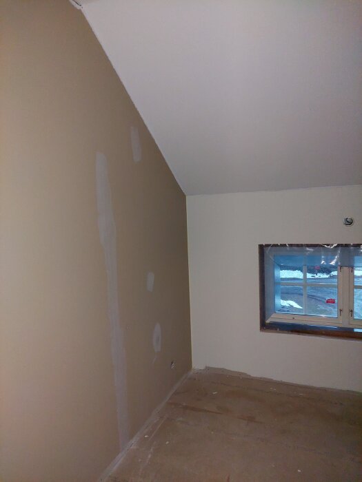 Rum under renovering med spacklad vägg, sned takvinkel och fönster som vetter mot snöig utsida. Ingen golvbeläggning.