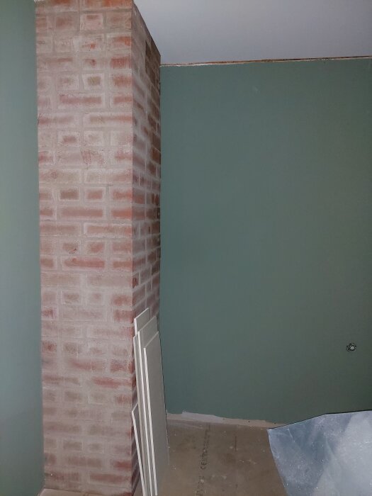 Tegelvägg inomhus bredvid målad vägg i grönt; plywoodskivor och plastfolie synliga på golvet.