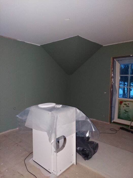 Rum under renovering med gröna väggar och vit maskin under plast. Fönster med utsikt och blommigt glas.