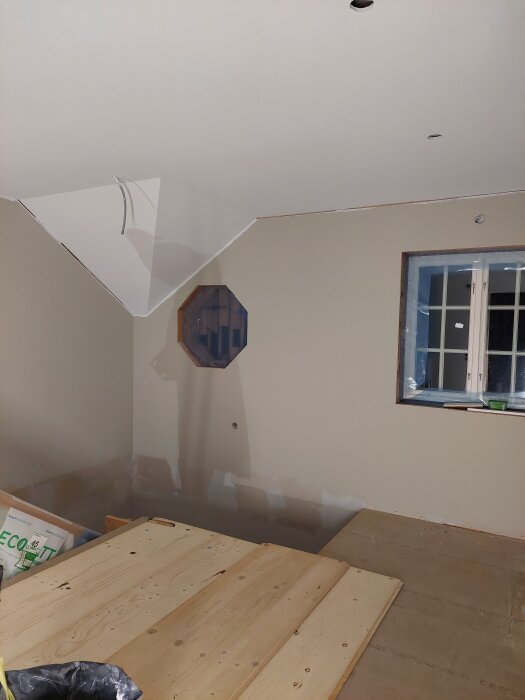 Renovering av rum med snedtak, träskivor på golvet, fönster och en octagonal öppning i väggen.