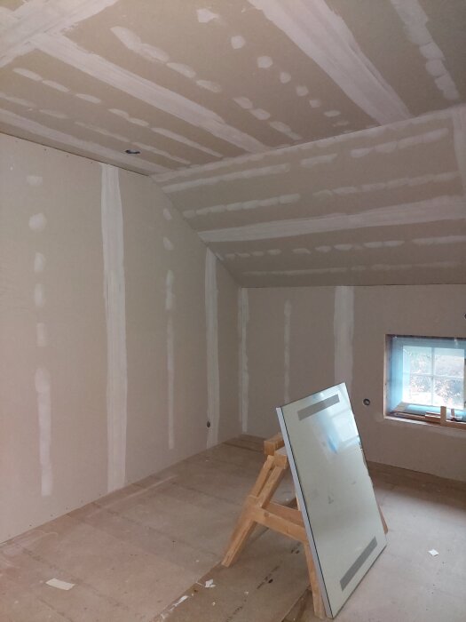 Ett rum under renovering med gipsväggar och ett tak, en stege och ett fönster. En skiva står lutad mot en bock.