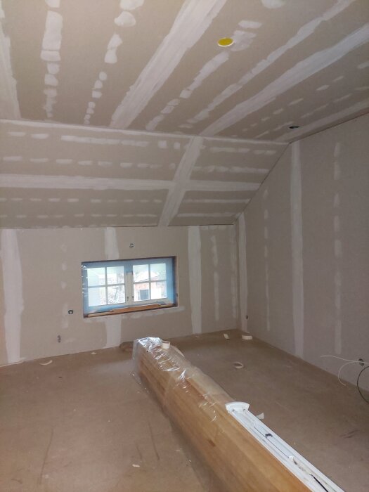 Ett rum under renovering med gipsskivor och spacklade skarvar. Ett paket golvmaterial ligger inplastat mitt på golvet, och ett fönster syns i bakgrunden.
