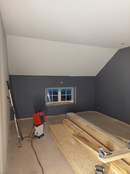 Byggprojekt i ett rum med mörkgrå väggar och snedtak, framträder arbetsredskap, staplade träpaneler och en värmefläkt på golvet.