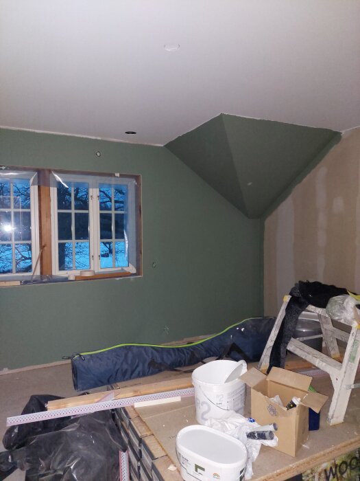 Rum under renovering med gröna väggar, en stege, målarburkar och en kartong på ett bord. Fönster med plastskydd och tegel syns utanför.