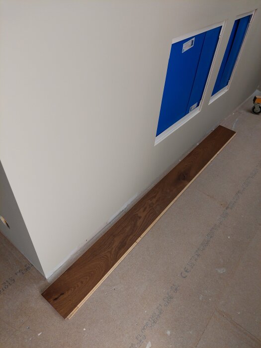 Del av ett rum med en ljus vägg, två blåa luckor och ett golv av brunt trä och spånskivor.