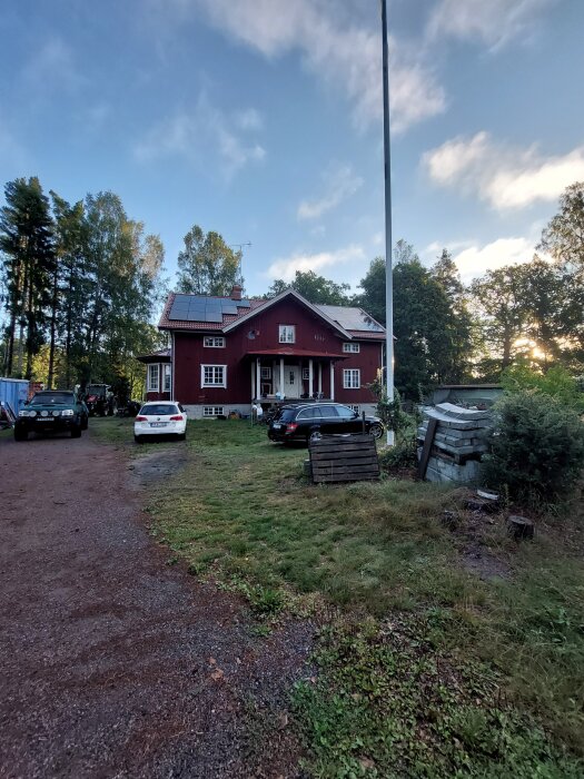 Rött trähus med vita knutar och solpaneler på taket, omgiven av bilar och grönska, i ett lantligt område vid solnedgång.