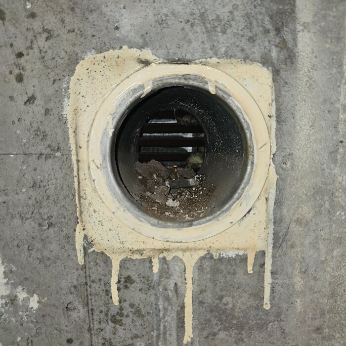 Rund ventilationsöppning i en betongvägg med misstänkta rester av gips och smuts inuti röret. Öppningen är cirka 120 mm i diameter.