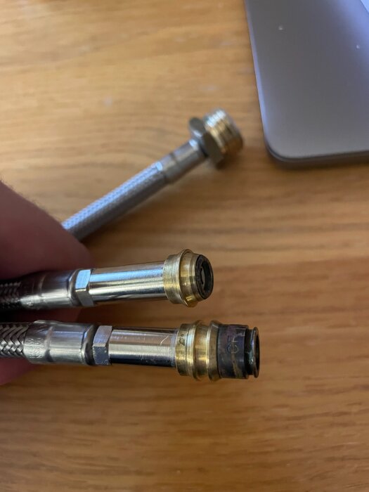 Två metallanslutningsrör med olika klämkopplingar, en oskarp bakgrund av trä och en bärbar dator syns delvis.