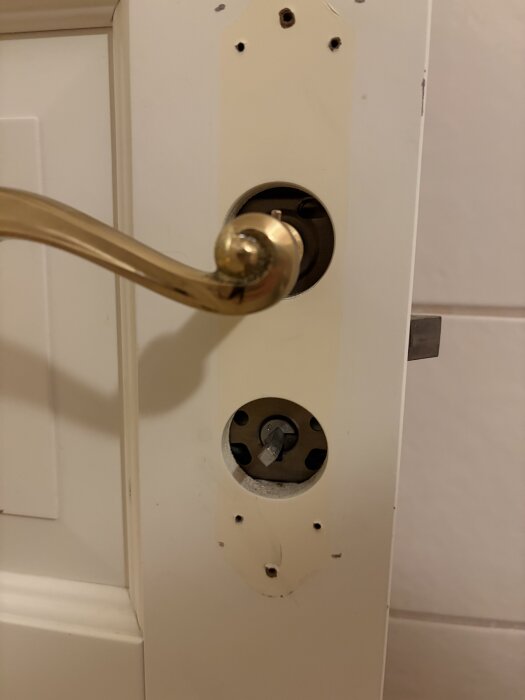 Gammalt dörrhandtag av mässing på en vit toalettdörr med borttaget vred, synliga hål runt vredets plats.