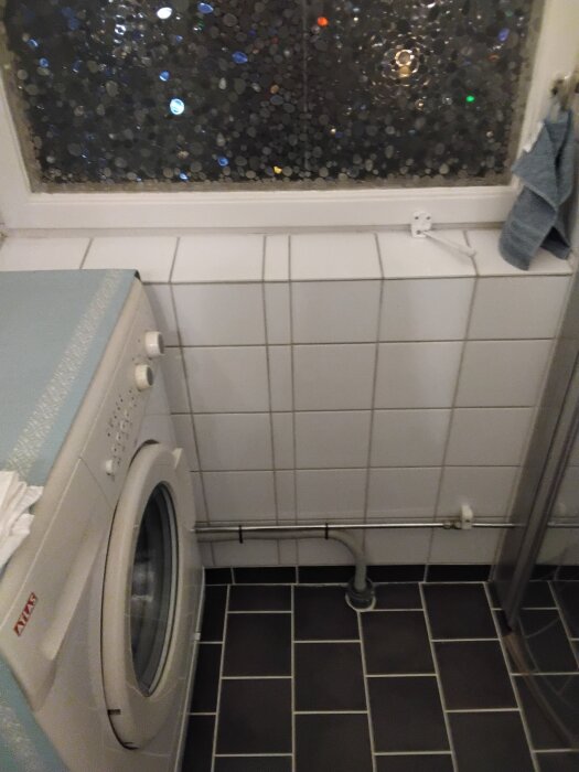 Tvättmaskin i ett kaklat badrum med mosaikmönstrat fönster, svart klinkergolv och synlig rördragning.