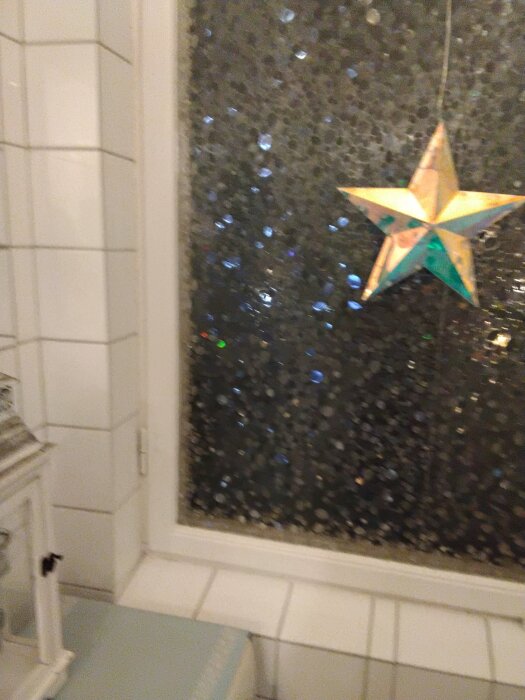 Stjärnformad ljusdekoration hänger i ett fönster med frostat glas, bredvid är vita kakelplattor synliga.