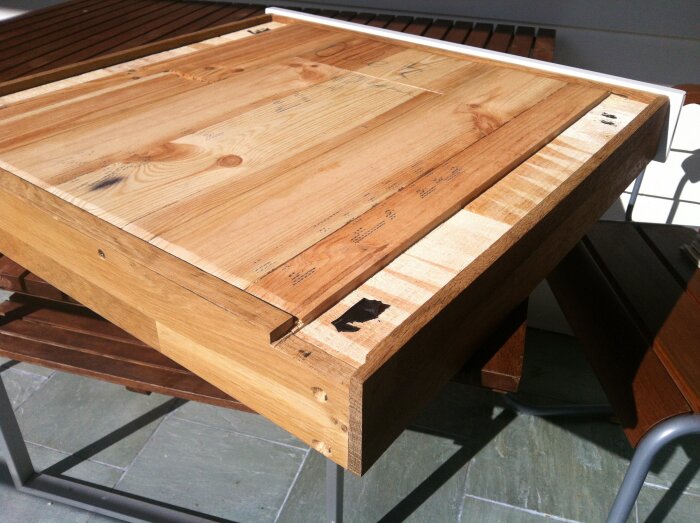 Träbordsskiva med synliga kvisthål och märken placerad på en utomhusbänk, väl lysande i solljus.