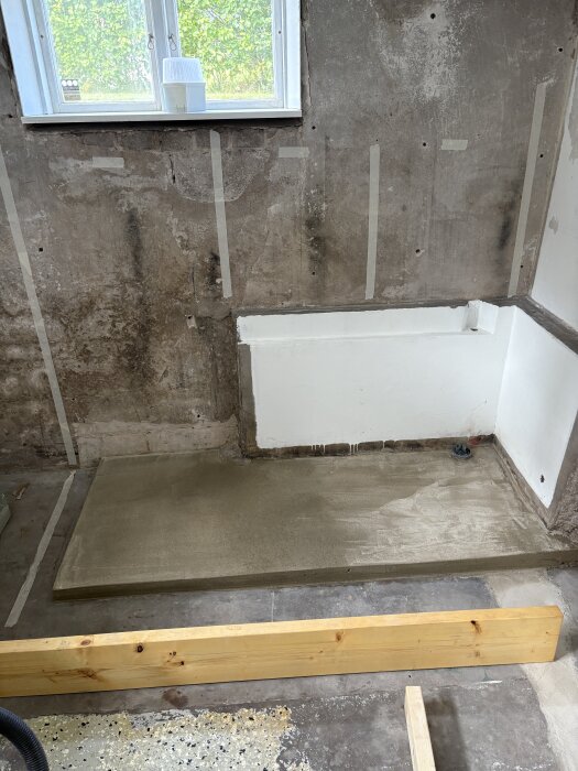 Betongklack under fönster med nygjuten yta och rör i hörnet. Halvfärdig tvättstuga med oputsade väggar och trägolvregel i förgrunden.