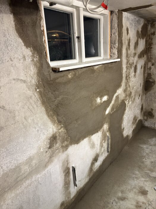 Betongvägg med nyligen lagade sprickor och nyputsad yta under ett fönster. Väggytan visar pågående renovering i tvättstugan.