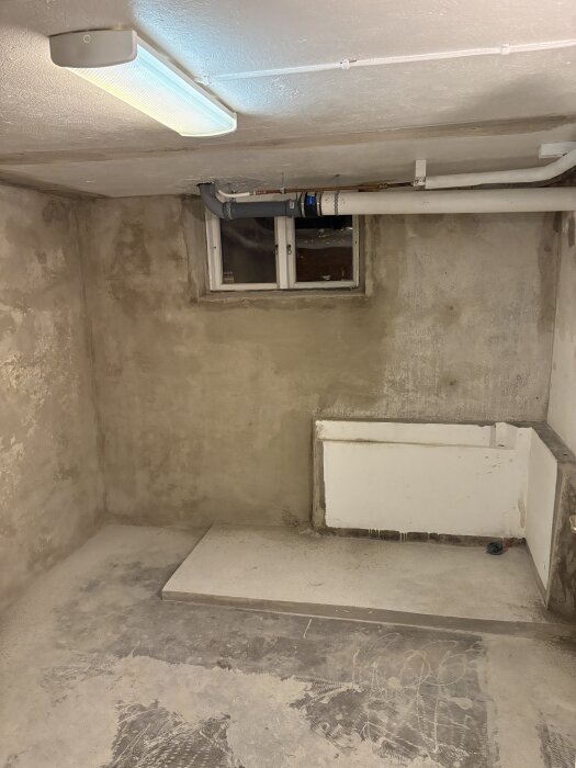 Orenoverad tvättstuga med synliga rör, gjuten betongklack, fönster och en lampa i taket. Golvet är delvis färdigbehandlat.